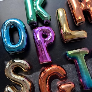 Puede incluir: Una colección de esculturas de letras metálicas de colores. Las letras, en varios colores como azul, verde, dorado y naranja, están dispuestas sobre una superficie oscura. El material brillante y reflectante crea una exhibición vibrante y llamativa.