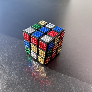 Könnte beinhalten: Ein schwarzer Rubik's Cube, dessen jede Seite mit bunten Strasssteinen bedeckt ist. Die Seiten sind rot, blau, grün, gelb, orange und weiß.
