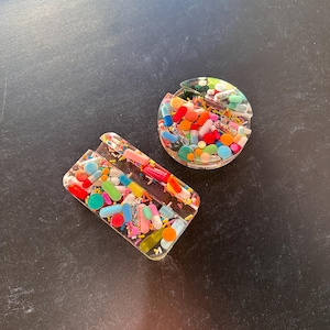 May include: Two clear resin coasters with a colorful assortment of pills and sprinkles embedded inside. The coasters are shaped like a circle and a rectangle.
