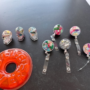 May include: An assortment of resin art pieces, including three clear skull-shaped objects filled with colorful beads, several badge reels with various colorful fillings, and a large orange resin piece with the word "LIVER" embedded.