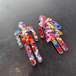 May include: Two clear resin figures shaped like stick people, filled with colorful pills, capsules, and glitter.