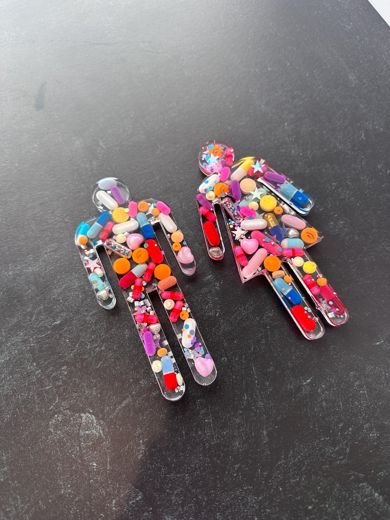 May include: Two clear resin figures shaped like stick people, filled with colorful pills, capsules, and glitter.