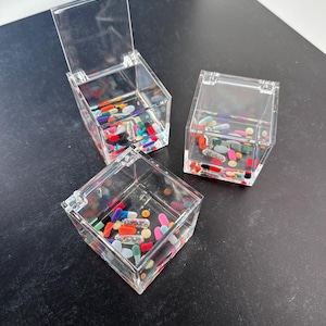 Peut inclure: Trois boîtes carrées en acrylique transparent remplies de pilules colorées et de paillettes. Les boîtes sont carrées et ont des couvercles. Les boîtes sont sur une surface noire.
