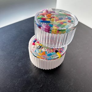 Può includere: Due contenitori di plastica trasparente con base bianca. I contenitori sono pieni di pillole e capsule colorate. I contenitori sono impilati uno sopra l'altro.