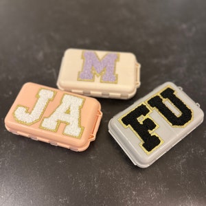 Può includere: Tre piccole scatole per pillole con coperchi di diversi colori e lettere in glitter dorato che compongono le parole "JA", "M" e "FU".