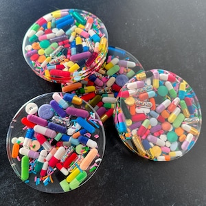 Puede incluir: Cuatro posavasos redondos de resina transparente con píldoras y chispas de colores incrustadas en el interior. Los posavasos tienen un diámetro de aproximadamente 10 cm.
