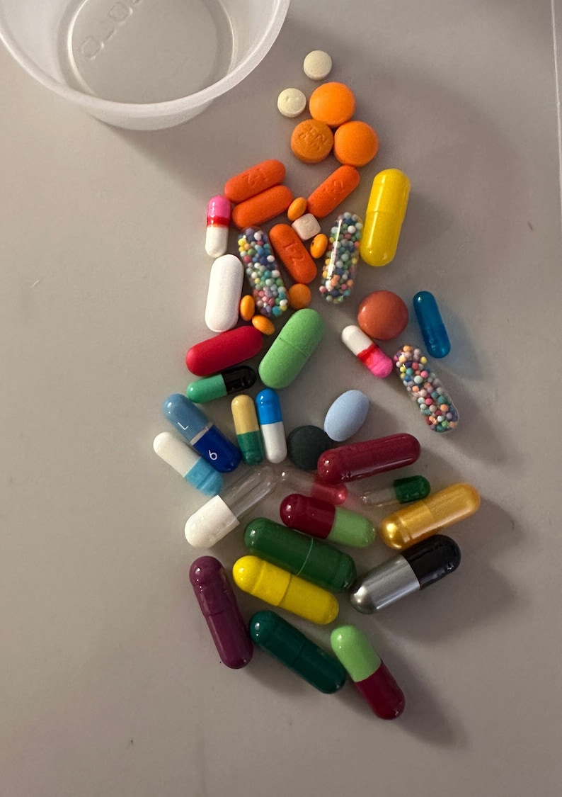 May include: Assorted pills and capsules in various colors and shapes, including orange, white, green, and blue. Some capsules are clear with colorful granules inside. A small white plastic cup is in the upper left corner.