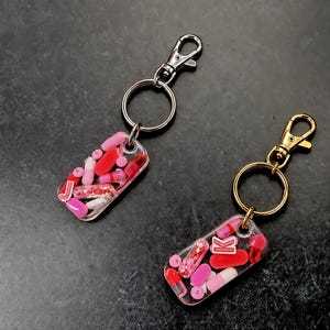 May include: Two rectangular resin keychains featuring embedded pink and red pill capsules and small beads. One keychain has a silver-tone metal lobster clasp and ring with the letter J, while the other has a gold-tone clasp and ring with the letter K.