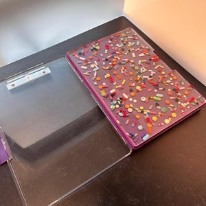 Könnte beinhalten: Ein durchsichtiges Clipboard aus Plastik mit einem violetten Innenraum. Das Clipboard ist geöffnet und der Innenraum ist mit bunten Pillen und Kapseln gefüllt.