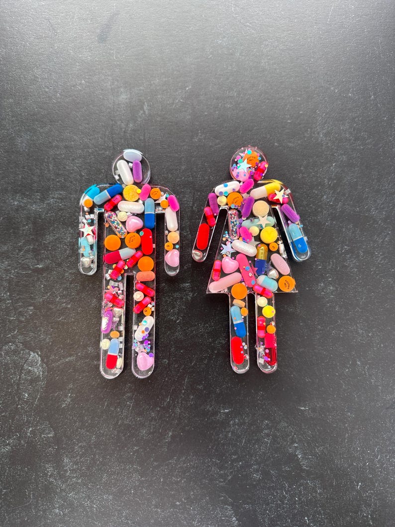 May include: Two clear resin figures shaped like people, filled with colorful pills and glitter. The figures are standing on a dark gray surface.