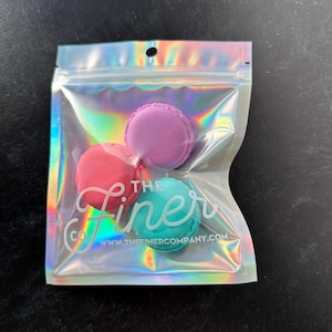 Puede incluir: Tres jaboneras de silicona con forma de macaron de colores diferentes en una bolsa transparente iridiscente. La bolsa tiene una etiqueta blanca con el texto "The Finer Co. www.thefinercompany.com".
