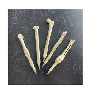 Peut inclure: Cinq stylos en forme d'os dans différents styles, dont un fémur, un humérus, une côte et une colonne vertébrale. Les stylos sont tous blancs et ont une texture osseuse réaliste.