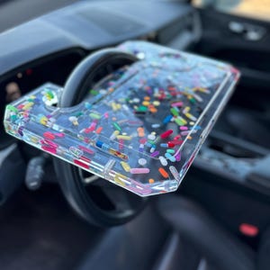 Peut inclure: Un plateau en plastique transparent avec un assortiment coloré de pilules incorporées à l'intérieur. Le plateau est fixé au volant d'une voiture.