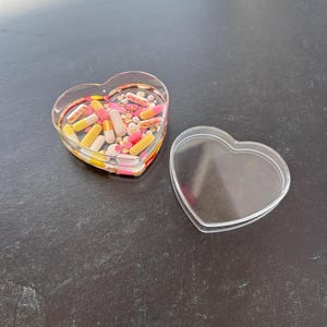 May include: A clear, heart-shaped container filled with various colorful pills and capsules. The container is open, with the lid placed beside it. The pills are in shades of yellow, pink, and white.