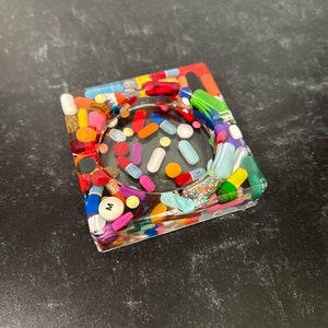 May include: A square, clear resin trinket dish filled with colorful pills and capsules. The dish has a raised rim and a smooth, glossy finish.