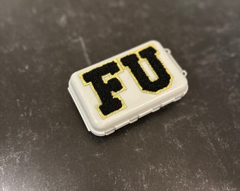 Puede incluir: Un estuche de almacenamiento rectangular de plástico blanco con las letras FU en tipografía negra de estilo universitario y un borde de purpurina dorada. El estuche tiene una tapa con bisagras y un mecanismo de cierre seguro en el lateral.