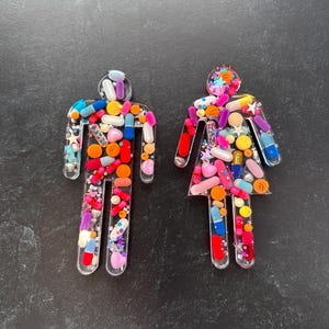 May include: Two clear resin figures shaped like people, filled with colorful pills and glitter. The figures are standing on a black surface.