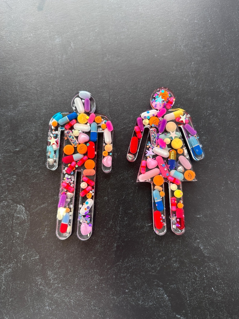 May include: Two clear resin figures shaped like people, filled with colorful pills and glitter. The figures are standing on a black surface.