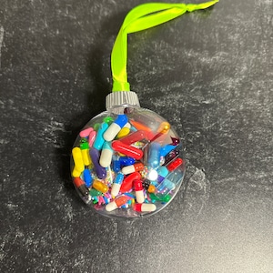Puede incluir: Una bola de Navidad transparente y redonda llena de cápsulas y chispas de colores. El adorno tiene una tapa plateada y una cinta verde brillante para colgar. Las cápsulas son de varios colores, incluyendo rojo, amarillo, azul y blanco.