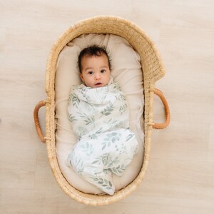 May include: A baby swaddled in a white and green leafy print swaddle blanket, lying in a woven basket.