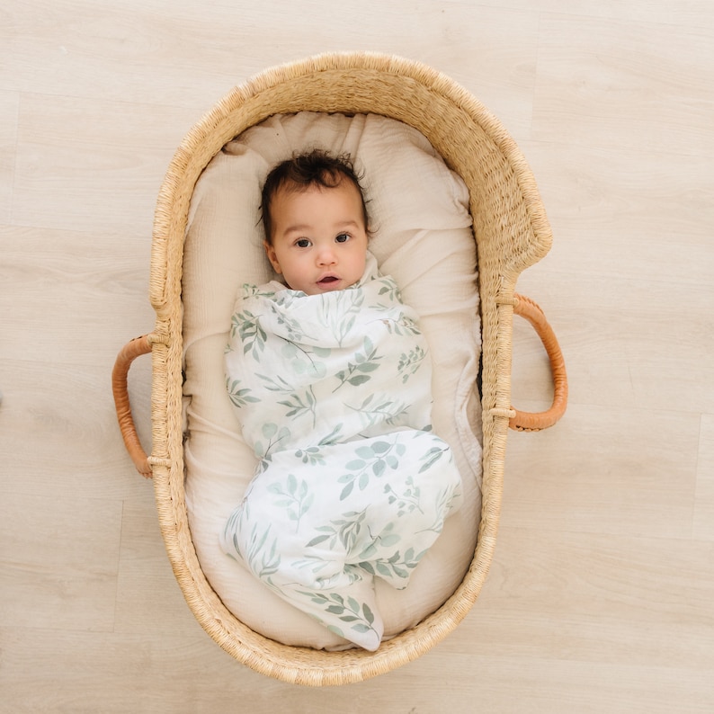 May include: A baby swaddled in a white and green leafy print swaddle blanket, lying in a woven basket.