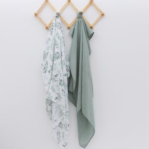 May include: Two muslin swaddles hanging on a wooden rack. One swaddle is white with a green leaf pattern, and the other is solid sage green.