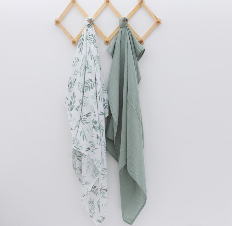 May include: Two muslin swaddles hanging on a wooden rack. One swaddle is white with a green leaf pattern, and the other is solid sage green.