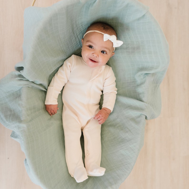 May include: A baby wearing a cream-colored long-sleeved onesie with a zipper down the front. The baby is lying on a light blue blanket and has a white bow in their hair.