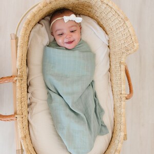 May include: A baby swaddled in a light blue muslin swaddle blanket, lying in a woven wicker bassinet. The baby is wearing a white bow headband.