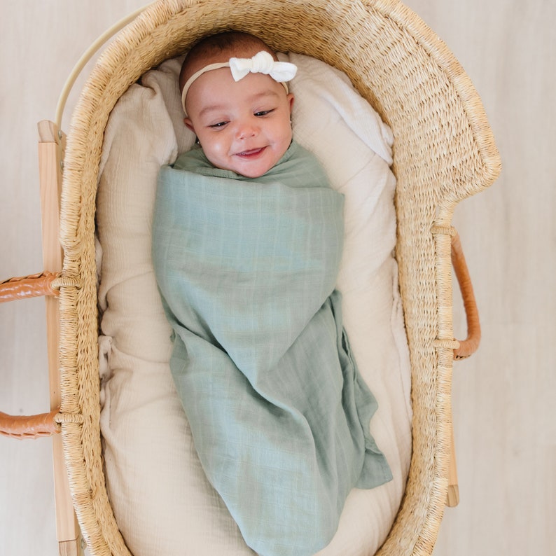 May include: A baby swaddled in a light blue muslin swaddle blanket, lying in a woven wicker bassinet. The baby is wearing a white bow headband.