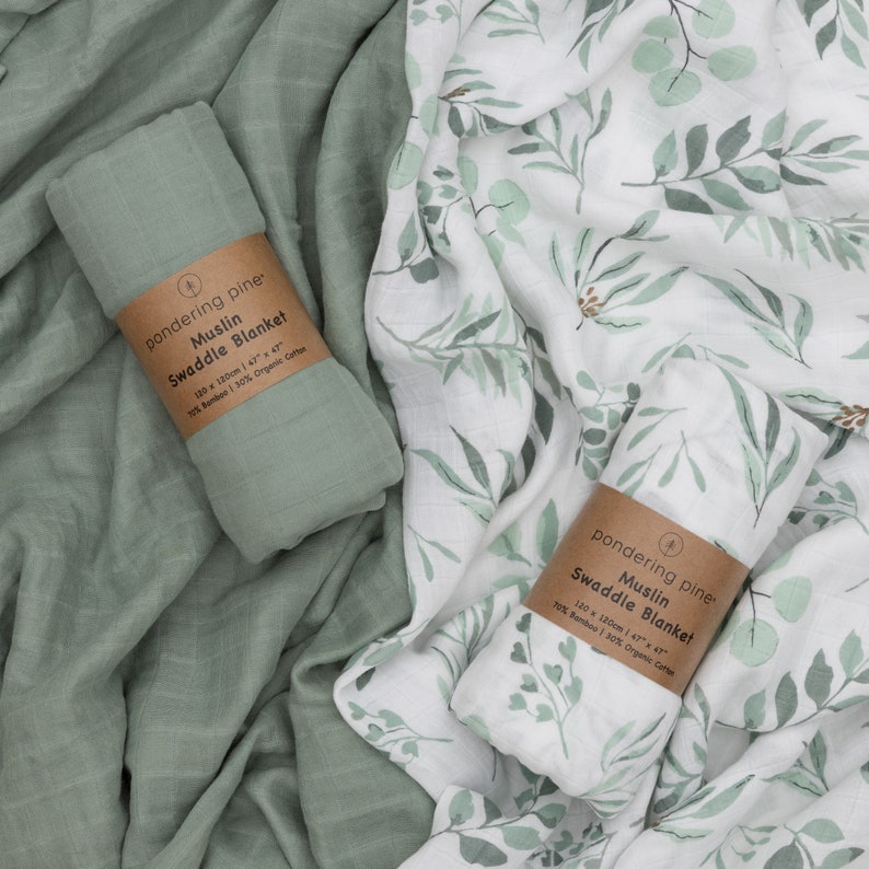 May include: Two green and white muslin swaddle blankets with a leafy pattern. The blankets are made of 77% bamboo and 30% organic cotton. The text on the label reads "pondering pine Muslin Swaddle Blanket 120 x 120cm 47" x 47".