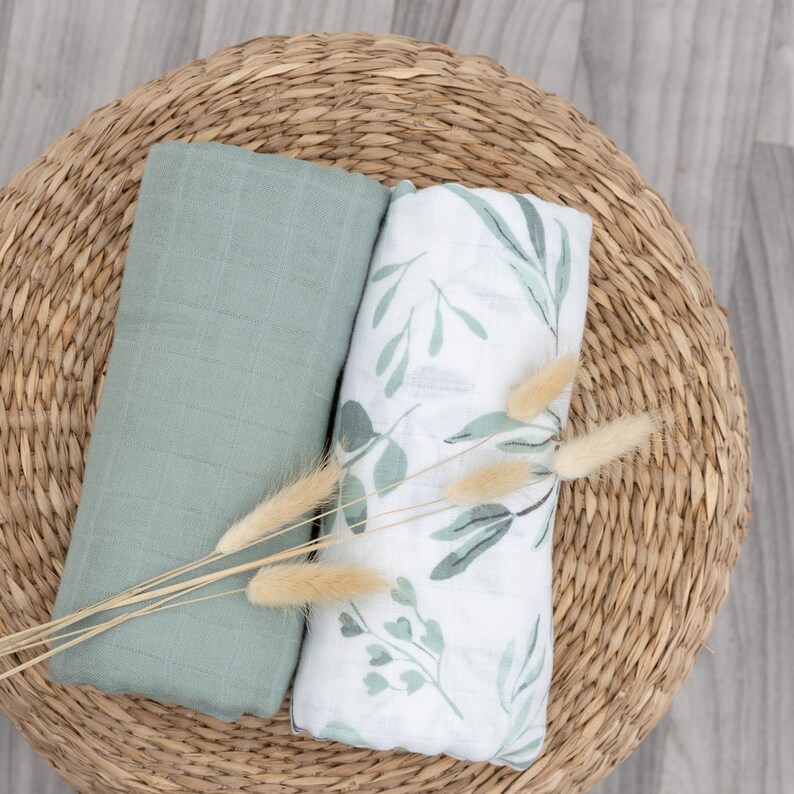 May include: Two soft, muslin swaddles, one green and one white with a leafy pattern. The swaddles are folded and placed on a woven basket.