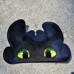 Può includere: Un adesivo nero raffigurante il volto di Sdentato di Dragon Trainer. L'adesivo ha grandi occhi verde brillante e un contorno bianco. L'adesivo è una forma ritagliata del volto del drago.