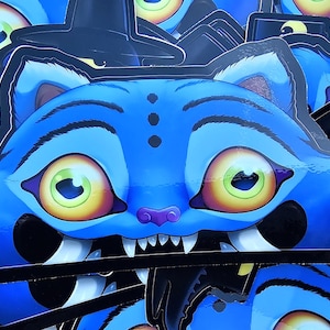 May include: Blue cat sticker with a witch hat. The cat has large yellow and green eyes, a purple nose, and sharp white teeth. The sticker is a Halloween-themed design.