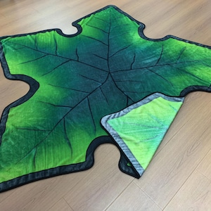 May include: A leaf-shaped blanket featuring a gradient of dark to light green colors with printed black vein details. The blanket has a black border trim and is folded over at one corner to reveal a lighter lime green underside.