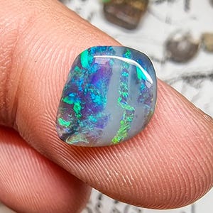 Puede incluir: Una piedra preciosa de ópalo pulido con una vibrante exhibición de colores, incluyendo azules, verdes y púrpuras. La piedra tiene una forma suave y redondeada y una apariencia translúcida. Otras piedras preciosas son visibles en el fondo.