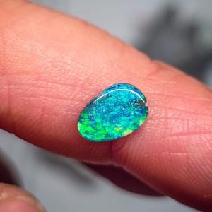 May include: A small, oval-shaped loose gemstone featuring a vibrant play of color with intense flashes of electric blue and bright green throughout its polished surface. The stone displays a smooth, rounded cabochon cut with a reflective finish.