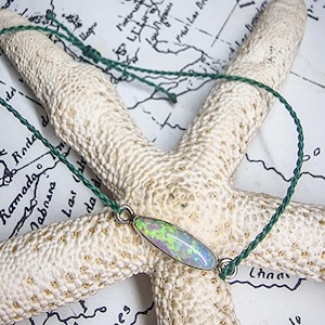 May include: A necklace with an oval opal pendant, displaying a spectrum of colours, set in a silver-tone bezel. The pendant hangs from a dark green cord. The necklace is displayed on a white starfish, against a vintage map background.
