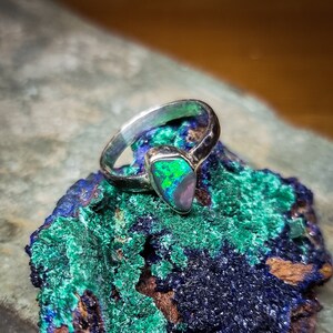 Puede incluir: Un anillo de plata con una piedra de ópalo verde y negra engastada en forma triangular. El anillo está apoyado sobre un trozo de malaquita azul y verde.