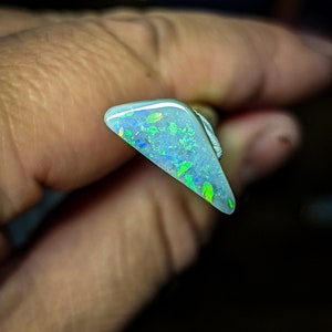 Puede incluir: Una pieza triangular de &oacute;palo con una base blanca y destellos iridiscentes verdes y azules.