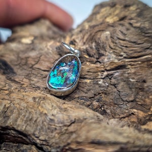 Peut inclure: Un pendentif en forme de goutte contenant une opale boulder sombre aux éclats irisés bleus et verts, serti dans une monture en argent poli avec une petite bélière circulaire en argent fixée sur le dessus pour le suspendre.