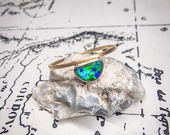 May include: A gold bracelet featuring a half-moon shaped opal with vibrant green and blue hues. The bracelet rests on a textured, light-colored stone, set against a backdrop of a map.