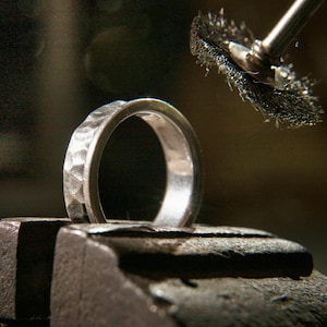 Könnte beinhalten: Ein silberner Ring mit einer strukturierten Oberfläche wird mit einer Drahtbürste poliert. Der Ring wird in einem Schraubstock aus Metall gehalten.