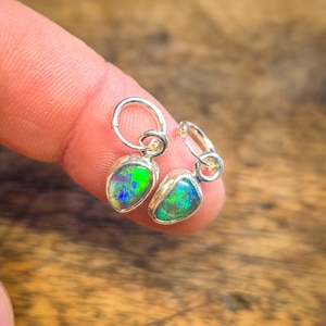 Peut inclure: Une paire de petites boucles d'oreilles créoles en argent. Chaque boucle d'oreille présente une pierre d'opale irisée en forme de goutte d'eau, aux teintes vertes et bleues vibrantes. Les boucles d'oreilles sont présentées sur un fond en bois flou.