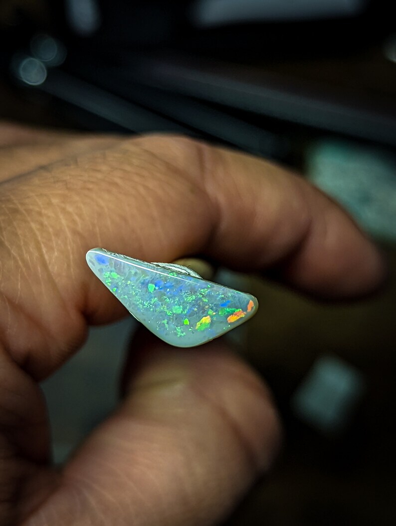 Puede incluir: Una pieza triangular de &oacute;palo con una base blanca y destellos iridiscentes verdes, azules y naranjas.