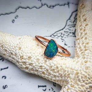 Puede incluir: Un delicado anillo de oro rosa con una piedra preciosa de ópalo de forma irregular única. El ópalo es de un color azul verdoso vibrante con destellos de color iridiscente.