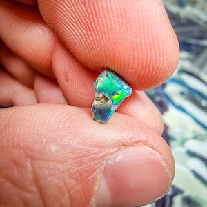 Puede incluir: Un pequeño fragmento de ópalo iridiscente, sostenido entre los dedos. El ópalo muestra destellos vibrantes de verde, azul y amarillo, con una superficie rugosa y texturizada. Los colores de la gema cambian con la luz, mostrando su belleza natural.