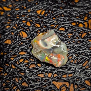 Può includere: Una gemma di opale grezza e traslucida con vibranti lampi di colore, tra cui rosso, giallo e verde. L'opale è posta su uno sfondo scuro e strutturato, che ne esalta la bellezza naturale. Questa gemma è un pezzo unico per gioielli o collezioni.