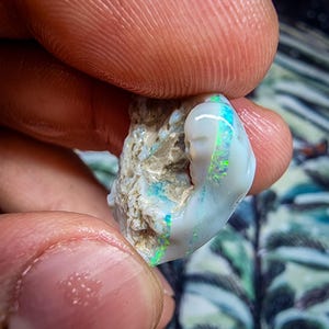 May include: A raw opal gemstone, held between fingers, with a creamy white base and flashes of green and blue iridescence. The uncut stone has a rough, natural texture. The opal's colours are vivid, with a play of light across its surface. The gemstone is a unique specimen.