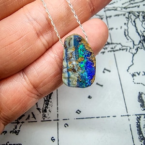Könnte beinhalten: Halskette mit Silberkette und Anhänger aus natürlichem Opal. Der Opal zeigt lebendige blaue, grüne und goldene Farben. Der Anhänger hat eine unregelmäßige Form und zeigt die einzigartigen Muster und Texturen des Steins.